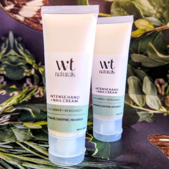 Two white tubes of Wriggly Tin Naturals Intense Hand & Nail Cream arranged on a dark surface surrounded by green leaves and dried botanicals.