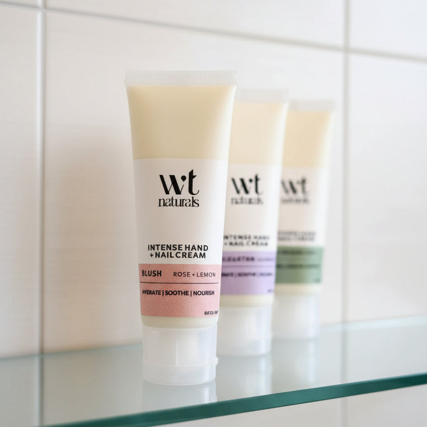 A tube of Wt Naturals Intense Hand + Nail Cream in the Blush Rose + Lemon scent, standing on a countertop with two more tubes blurred in the background.