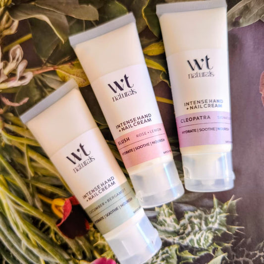 Three tubes of WT Naturals hand and nail creams arranged diagonally on a dark surface surrounded by dried botanical leaves and flowers. The tubes are labeled Intense Hand Nail Cream in Rose+Lemon, Intense Hand Nail Cream in Cucumber + Bergamot, and Intense Hand Nail Cream in Cleopatra.